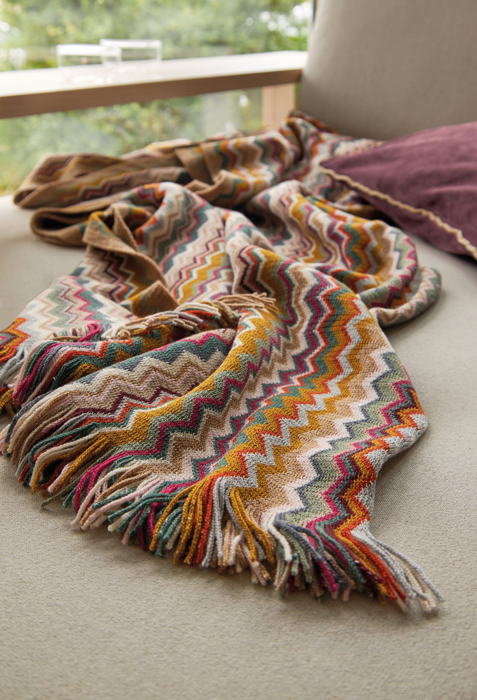 Bunte Zickzack-Decke und lila Kissen auf hellem Sofa, schaffen eine gemütliche Atmosphäre.