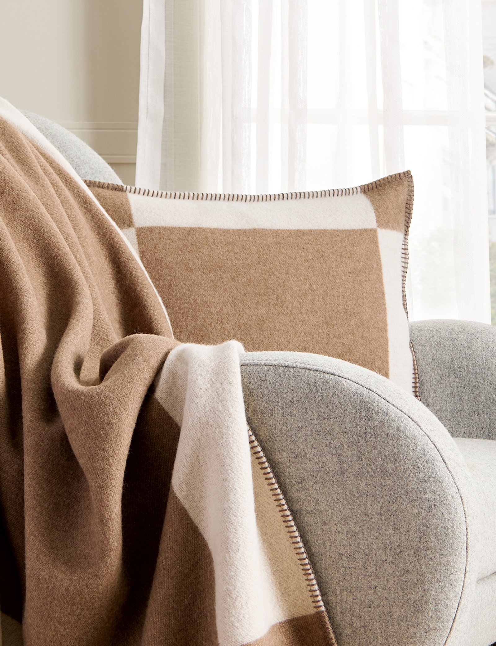 Gemütliches Wohnzimmer mit grauem Sessel, kariertem Kissen und Decke, sanft beleuchtet durch Tageslicht.