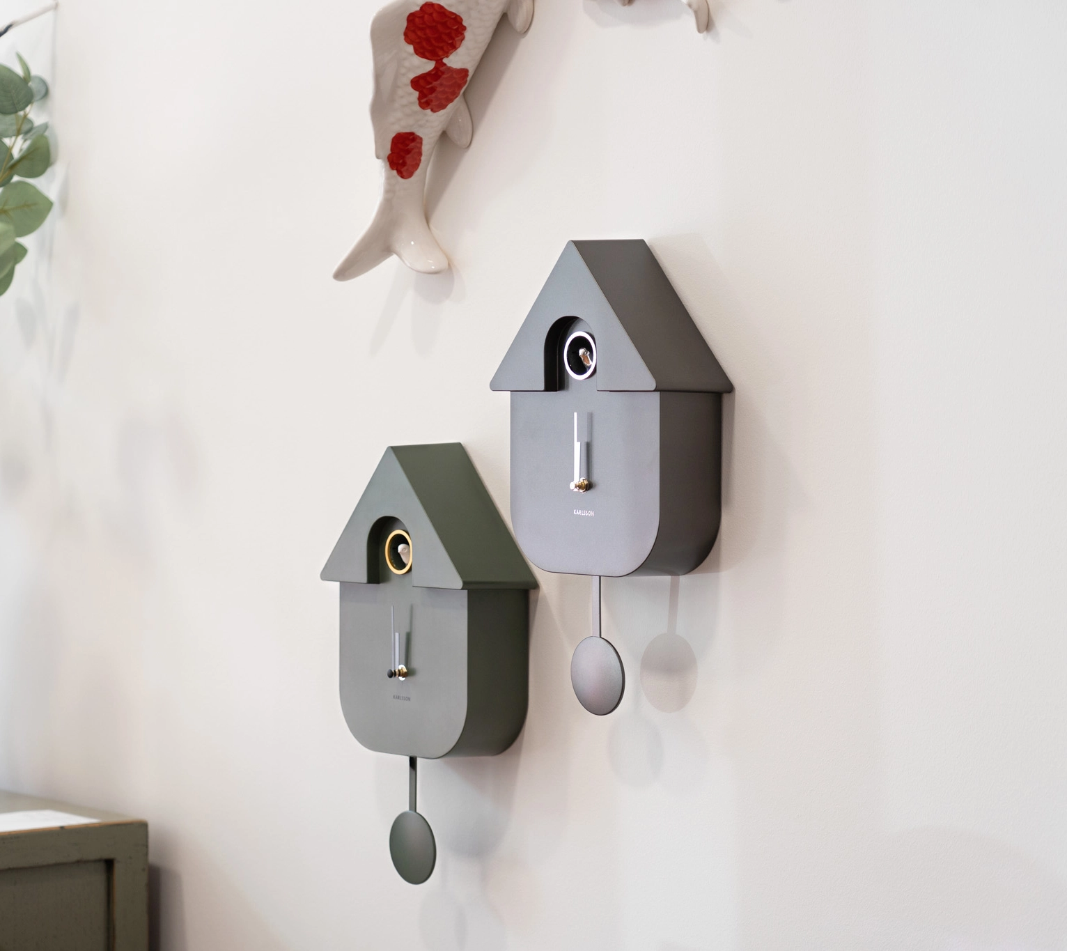 Zwei moderne Kuckucksuhren und ein dekorativer Keramikfisch hängen an einer hellen Wand.