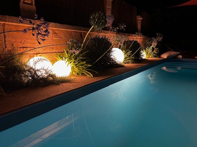 Ein beleuchteter Pool in einem nächtlichen Garten, umgeben von Pflanzen und warmen Kugelleuchten.