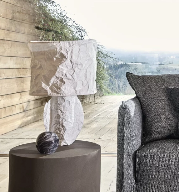 Modernes Wohnzimmer mit grauem Sofa, zerknitterter weißer Lampe auf rundem Tisch, Blick auf Natur.