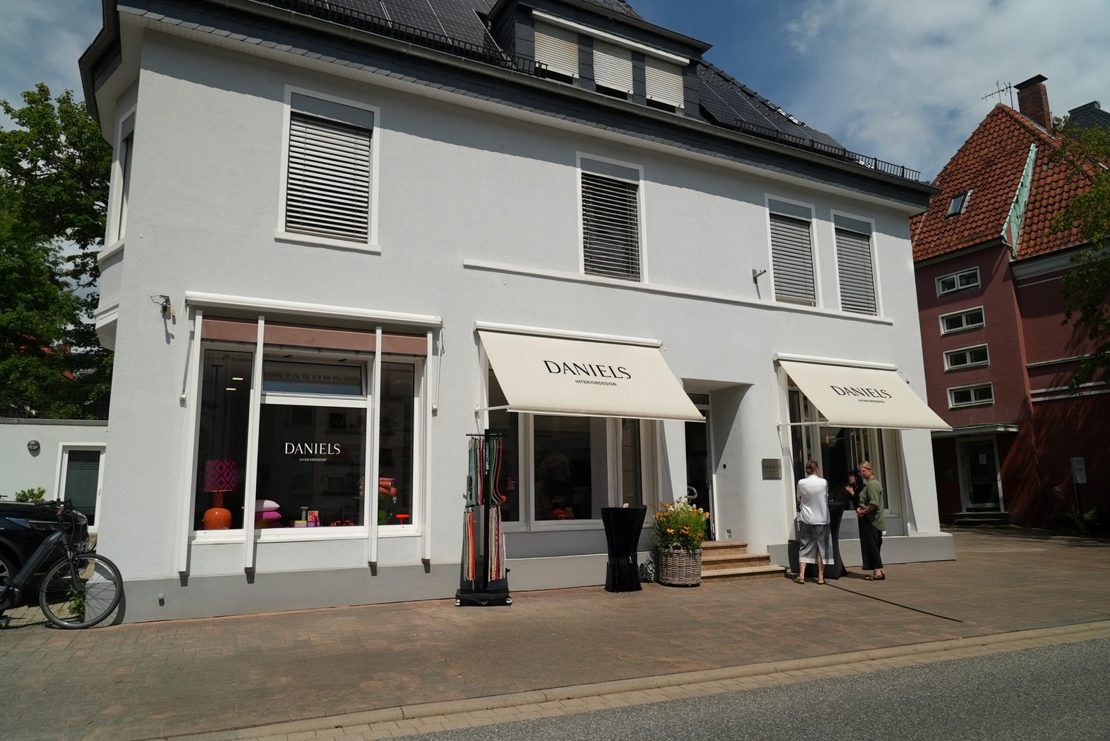 Zweistöckiges Geschäft "DANIELS" mit weißen Wänden, großen Fenstern und zwei Personen davor.